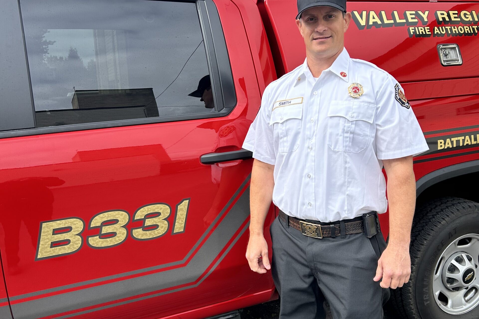 Newly promoted Battalion Chief Guy Smith, in uniform, stands beside a red fire department vehicle labeled