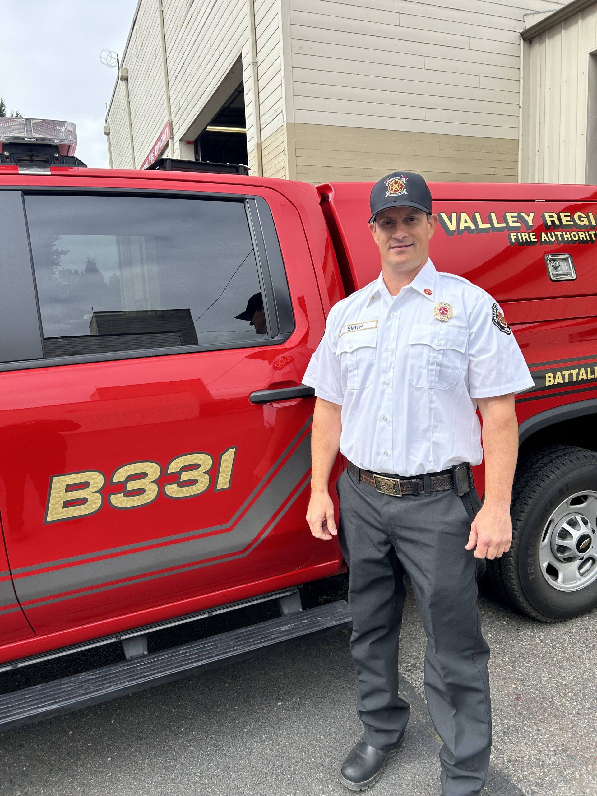 Newly promoted Battalion Chief Guy Smith, in uniform, stands beside a red fire department vehicle labeled