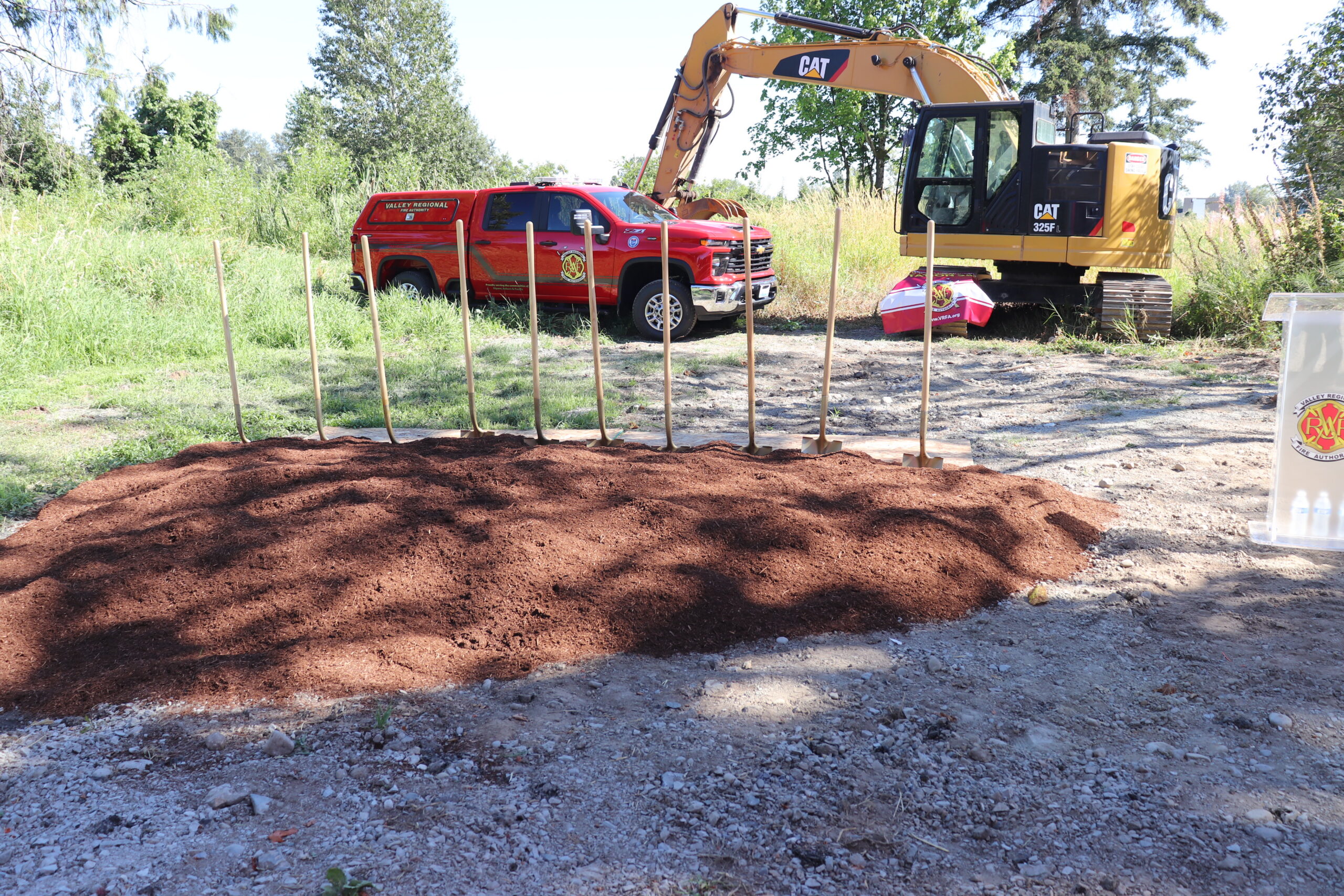 A row of shovels stands in front of a fresh dirt mound, with a red truck and yellow excavator parked nearby on a sunny day—signaling progress as part of the August 2026 Capital Facilities Update.