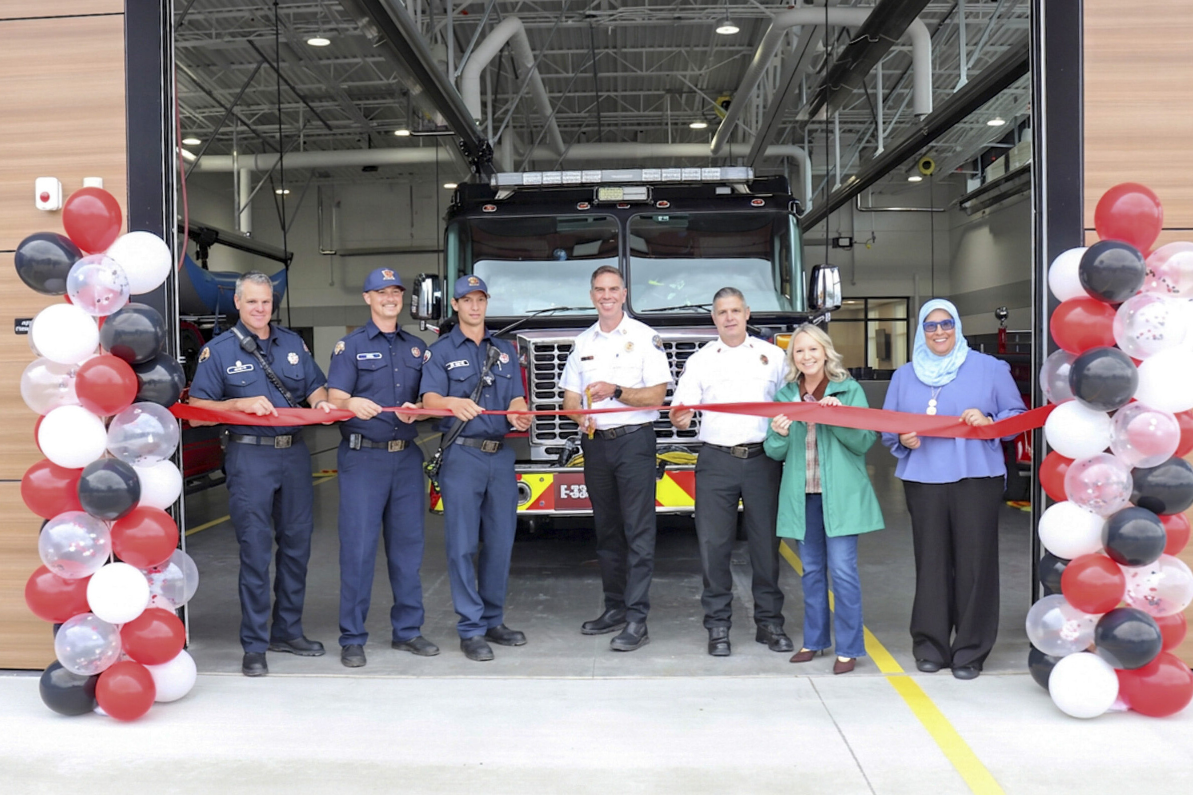 Seven people stand in front of a fire truck holding a red ribbon for a ceremony at the Open House of Station 36, flanked by columns of black, white, and red balloons inside the fire station.