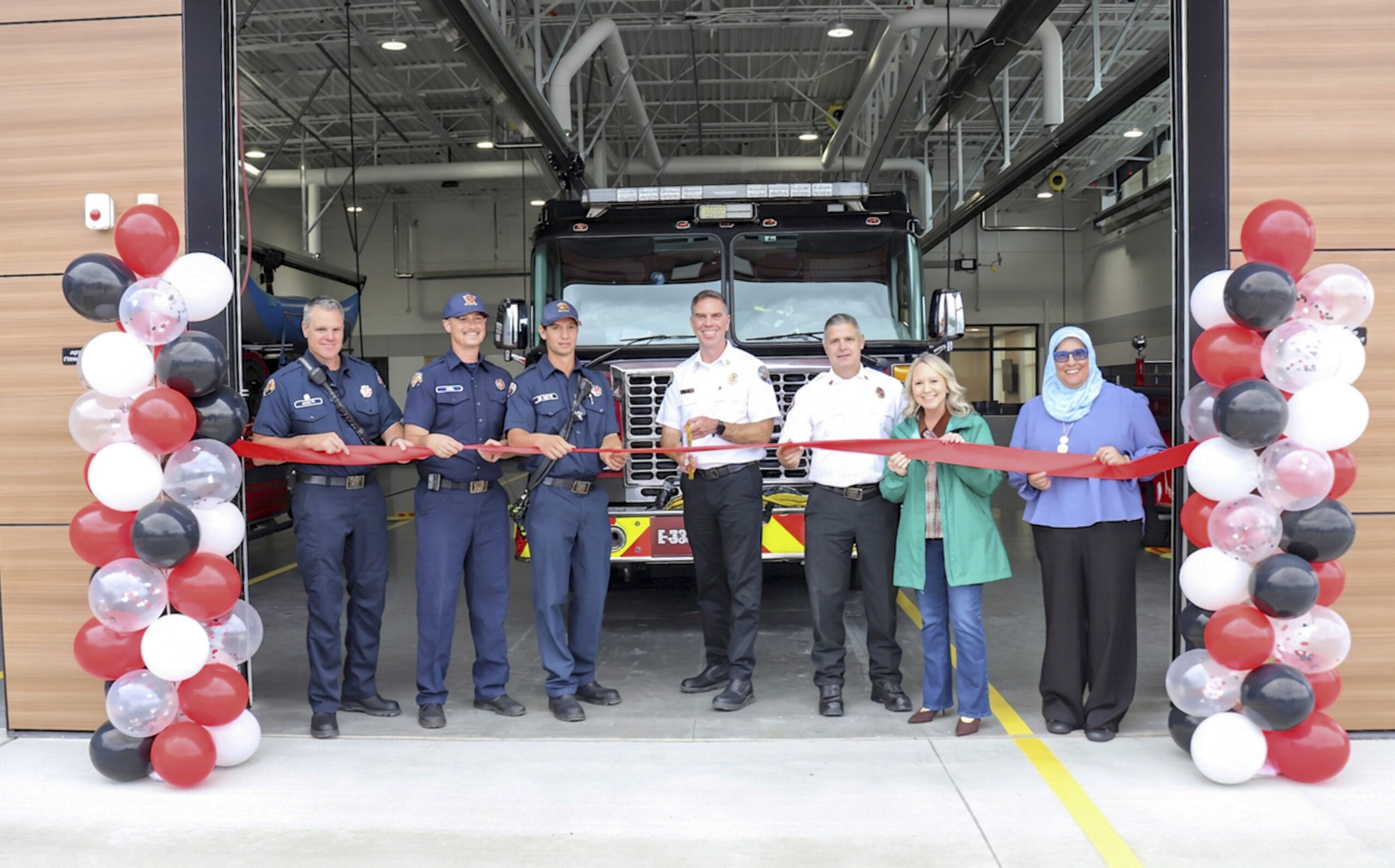 Seven people stand in front of a fire truck holding a red ribbon for a ceremony at the Open House of Station 36, flanked by columns of black, white, and red balloons inside the fire station.