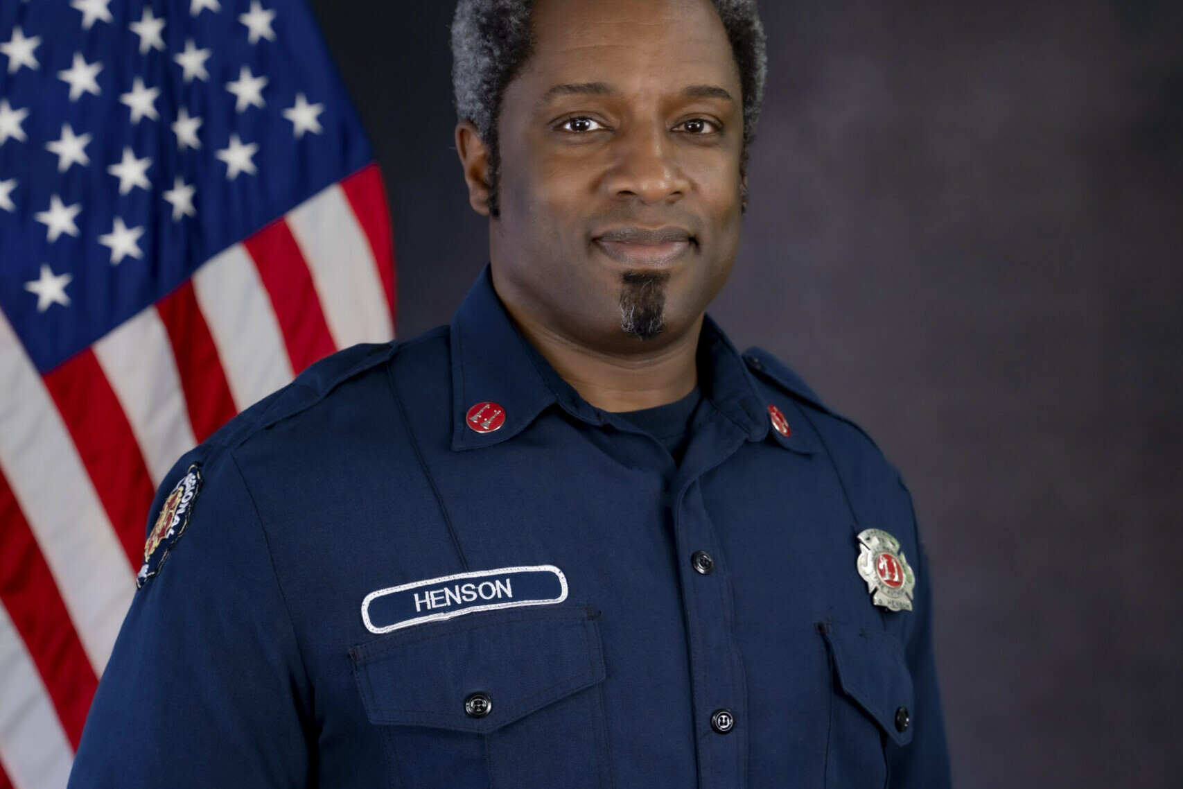 Captain Gervase Henson, a firefighter in uniform, stands in front of an American flag, looking at the camera with a neutral expression—a powerful image capturing his years of dedicated service as he approaches retirement.