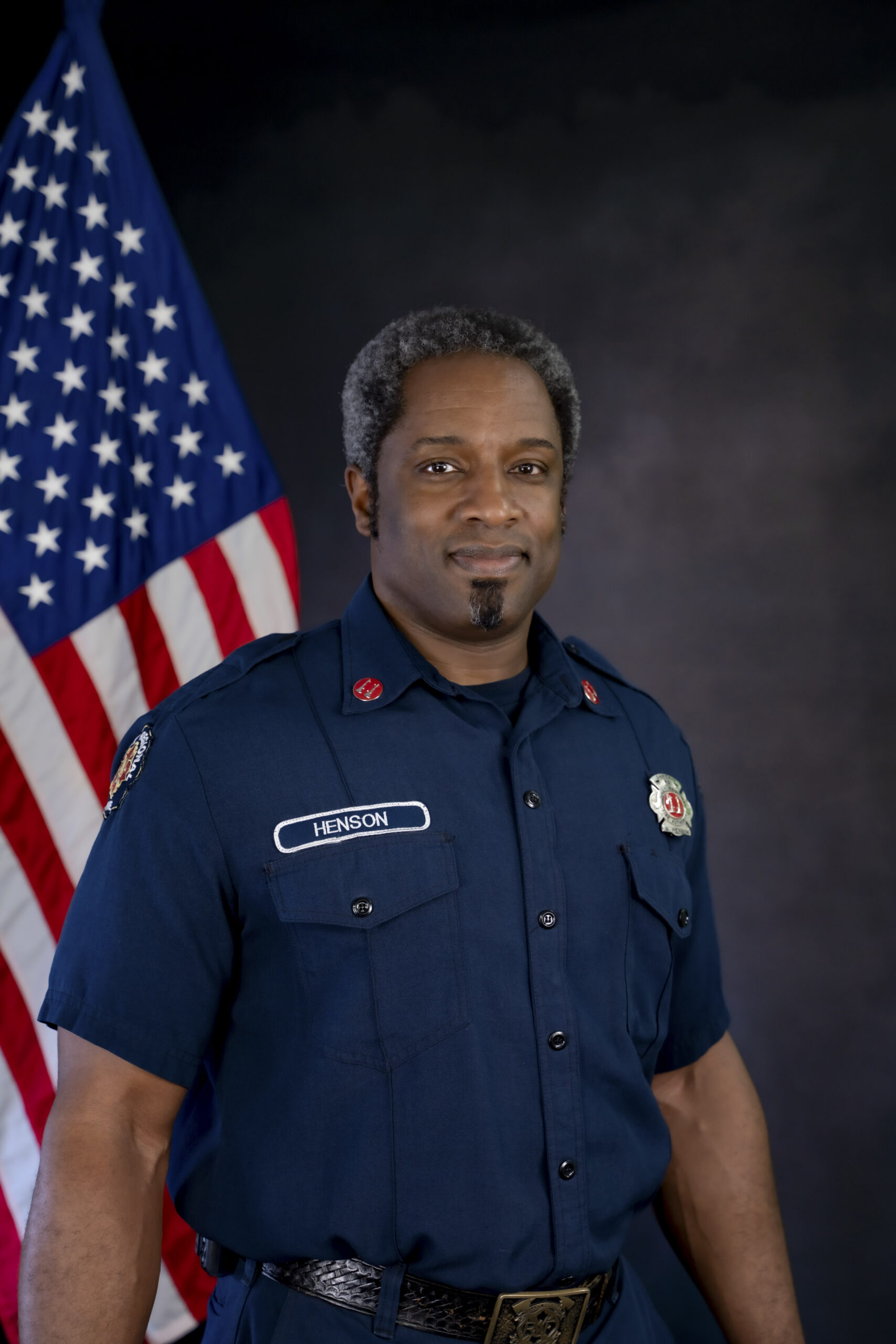 Captain Gervase Henson, a firefighter in uniform, stands in front of an American flag, looking at the camera with a neutral expression&mdash;a powerful image capturing his years of dedicated service as he approaches retirement.